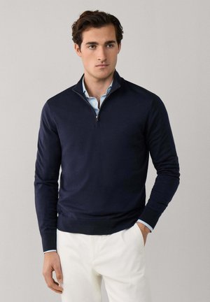 Hombre con suéter azul marino con cremallera en el cuello sobre una camisa azul claro y pantalones blancos, de pie frente a un fondo gris liso.
