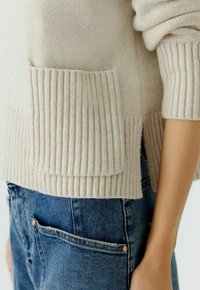 Beiger Strickpullover mit geripptem Saum und Seitentasche, kombiniert mit blauen Jeans. Die Textur ist weich mit sichtbaren Strickmustern.