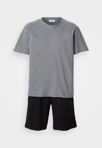 Απλή γκρι κοντομάνικη μπλούζα με στρογγυλή λαιμόκοψη συνδυασμένη με μαύρο μονόχρωμο σορτς μέχρι το γόνατο, σε λευκό φόντο.