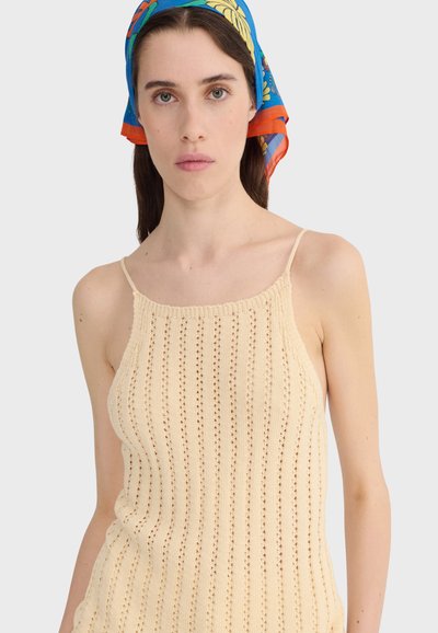 Femme aux longs cheveux bruns portant un foulard à fleurs bleu, orange et jaune, et un haut beige en tricot sans manches avec de fines bretelles.
