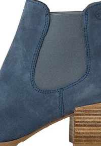 Blå mocka ankelskor med elastiska sidopaneler, träklossklack och kontrasterande sömmar. Texturerat material ger visuell intresse.
