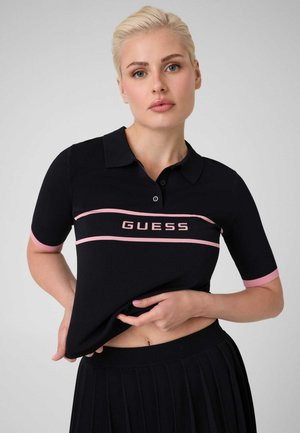 Femme aux cheveux blonds porte un haut noir à manches courtes avec bordure rose et texte "GUESS", soulevant légèrement l'ourlet pour révéler sa taille.