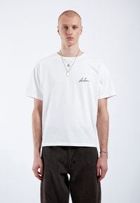 T-shirt en coton blanc avec un col rond et des manches courtes, arborant du texte manuscrit noir sur la poitrine gauche. Porté avec un pantalon en denim noir.