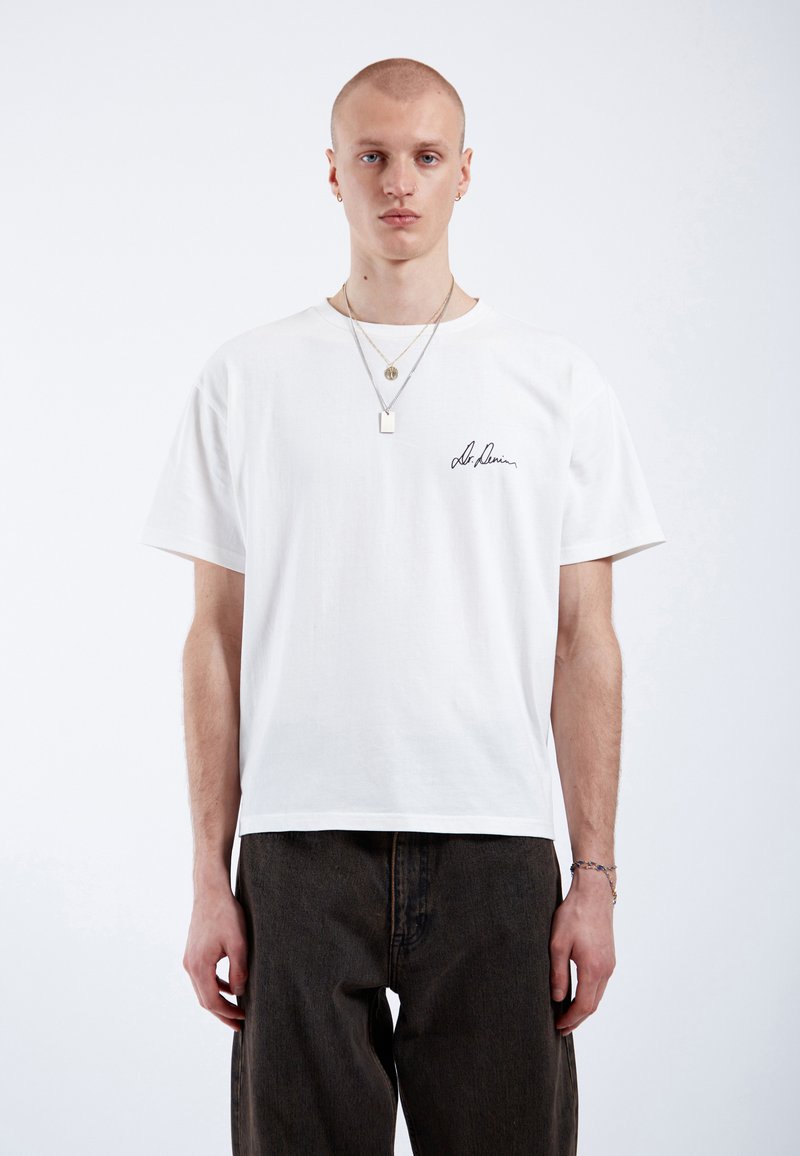 T-shirt en coton blanc avec un col rond et des manches courtes, arborant du texte manuscrit noir sur la poitrine gauche. Porté avec un pantalon en denim noir.