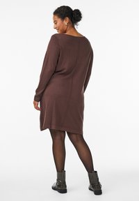 Bruine gebreide jurk met lange mouwen, ronde halslijn en een rechte snit die boven de knie valt. Heeft een centrale achternaad en geribbelde boorden.