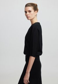 Pull en maille noir avec des manches trois-quarts et un col rond, associé à une jupe noire assortie, affichant une silhouette élégante.