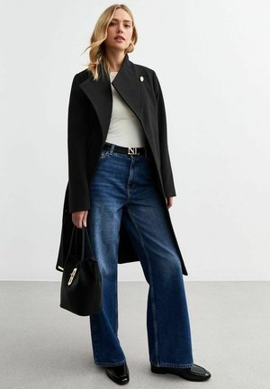 Manteau noir long avec un col pointu, porté sur un haut blanc. Associé à un jean bleu à jambes larges et un sac à main noir. Chaussures noires brillantes.