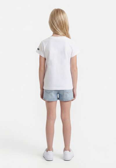 Svetlolasi otrok, ki stoji obrnjeno stran, nosi preprosto belo majico s kratkimi rokavi, svetlo modro jeans kratke hlače in bele superge na enobarvnem belem ozadju.
