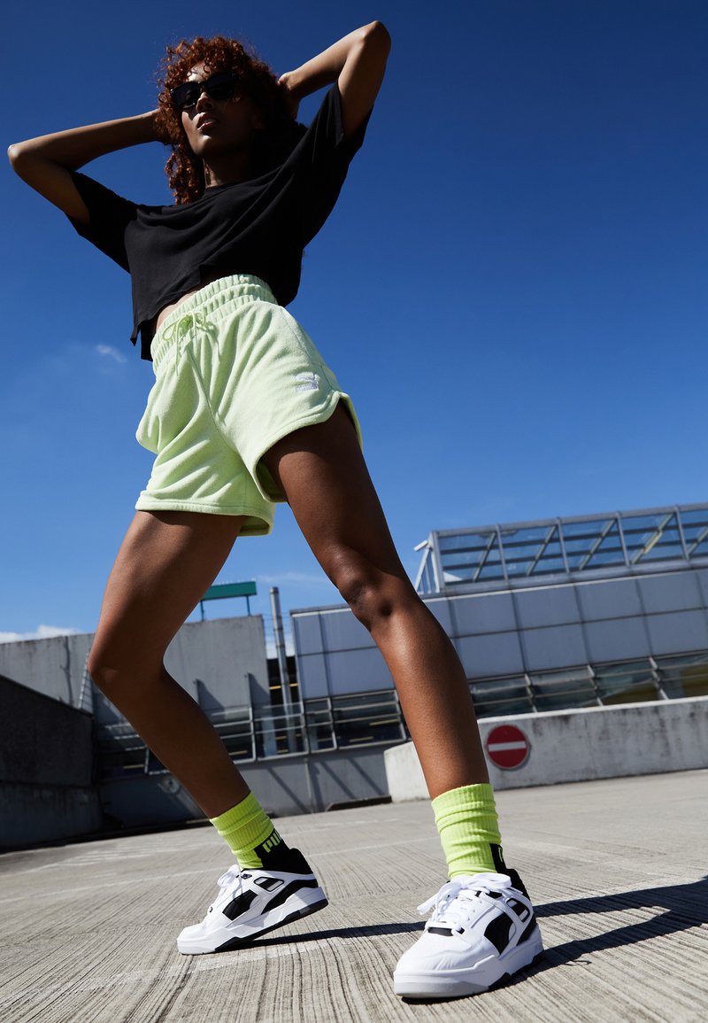 Svart avkortet T-skjorte, lysegrønne shorts med elastisk strikk i livet, neongrønne sokker og svart-hvite joggesko mot en blå himmel.