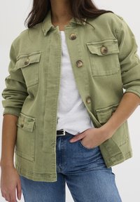 Olive green canvas jacket with a button-up front, two chest pockets, and rolled sleeves, paired with a white shirt and blue jeans.