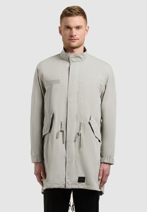 Homme portant un long manteau gris clair avec un col haut, des poches avant à rabats et une taille à cordon, debout devant un fond uni.