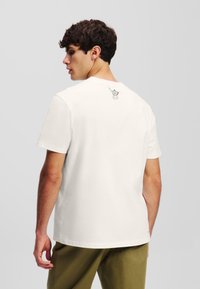 Hvid bomulds t-shirt med korte ærmer, med et minimalistisk sort grafik på ryggen. Modellen har olivengrønne bukser på og står sidelæns.