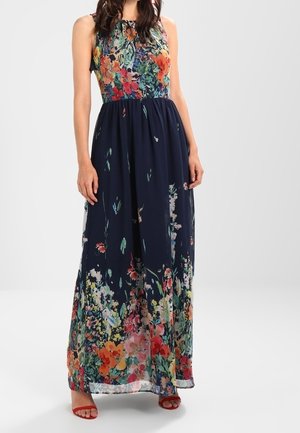 Sukienka maxi bez rękawów z dopasowanym kwiecistym gorsetem i granatową spódnicą z żywym, wielokolorowym kwiatowym wzorem, noszona z czerwonymi sandałami na otwartym palcu.