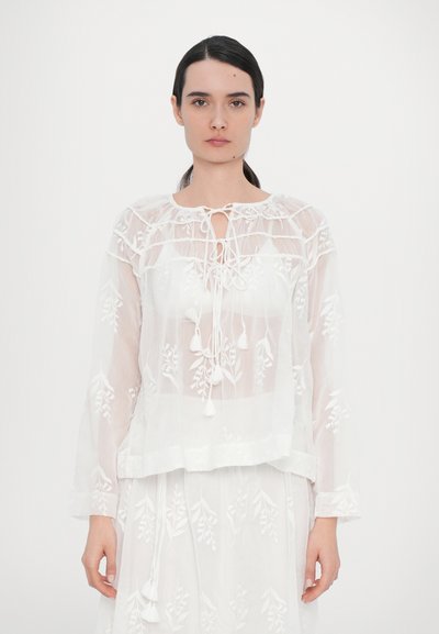 Blouse blanche brodée avec un col à cordon, présentant des motifs floraux, des manches longues et des accents de franges. Tissu léger et transparent.