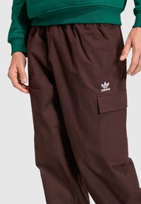 Pantalones cargo marrones con tela texturizada, cintura elástica y bolsillo lateral; logotipo de Adidas blanco en el muslo izquierdo.