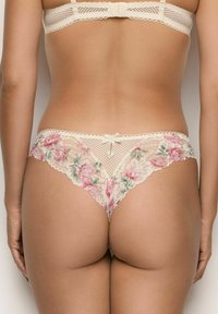 Vrouw in beige kanten lingerie met roze bloemenpatronen, van achteren gezien tegen een effen achtergrond.