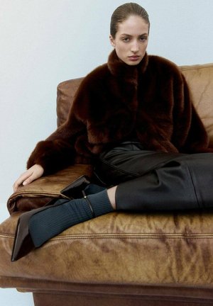 Brown faux fur cropped jacket, black leather pants, and textured dark teal socks. The model sits on a brown leather couch.