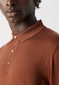 Mann in einem rostfarbenen polohemd mit Knöpfen und kleinem Kragen, sichtbarem Ohrstecker und Nahaufnahme von Lippen und Halsbereich.