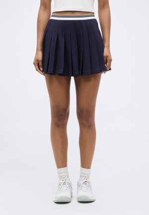 Žena v námornícky modrej plisovanej tenisovej sukni s bielym a čierno pruhovaným pásom, bielymi ponožkami a bielymi športovými topánkami.