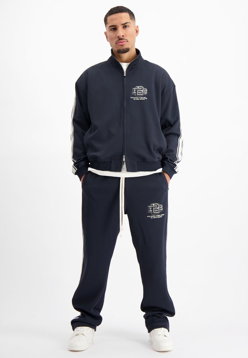 Marineblauwe trainingsuit met witte accenten, bestaande uit een jack met rits en een broek met trekkoord. De stof lijkt zacht, met geborduurd logo op beide stukken.