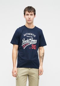 Tricou din bumbac de culoare bleumarin, cu guler rotund și mâneci scurte. Prezintă grafica de text alb și roșu, inclusiv "Jack & Jones" și "90."