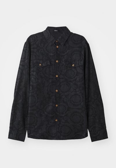 Chemise noire à manches longues avec un motif texturé, deux poches poitrine et des boutons marron sur le devant à boutons. Design avec col.
