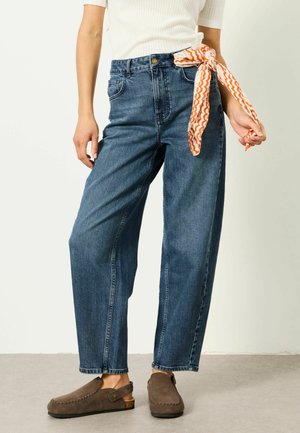Person trägt hoch taillierte blaue Jeans mit einem orange-weiß gemusterten Schal, der an der Gürtelschlaufe befestigt ist, braune Slip-On-Schuhe und ein weißes Oberteil.