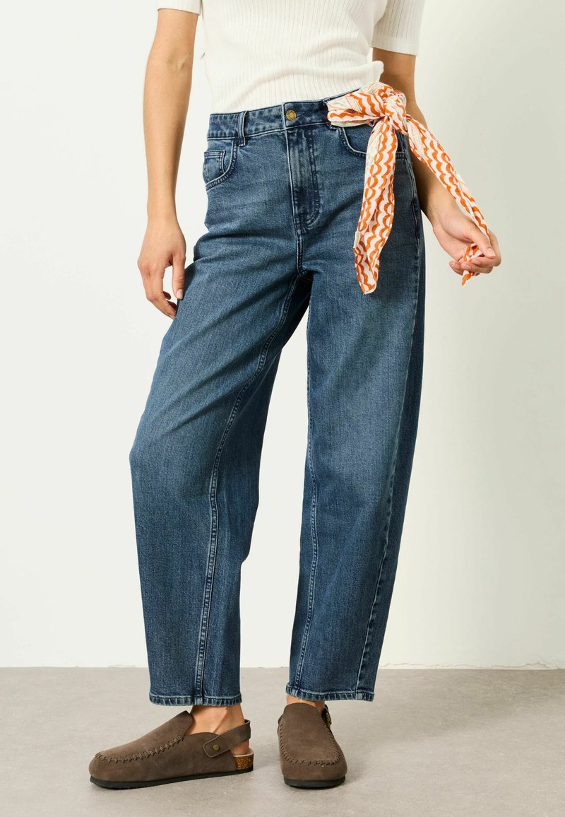 Persona che indossa jeans a vita alta blu con una sciarpa arancione e bianca con motivo annodata all'asola della cintura, scarpe marroni slip-on e una maglietta bianca.