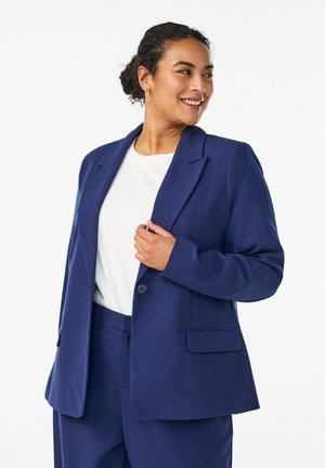 Lächelnde Frau mit lockigem Haar, gekleidet in einen dunkelblauen Anzug und ein weißes Hemd, hält das Revers ihrer Jacke und blickt seitlich vor weißem Hintergrund.