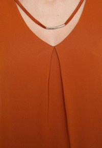 Blusa di colore ruggine con scollo a V, caratterizzata da una piega al centro e hardware curvo di tonalità argentata sul colletto. Texture liscia.