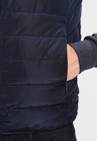Marineblaue Steppweste mit genoppter Optik und Seitentasche, aus glänzendem Stoff gefertigt. Graue Strickbündchen sind unter der Weste sichtbar.