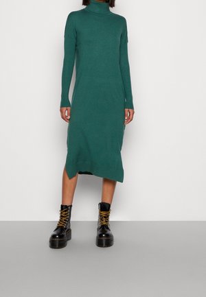 Person trägt ein langärmeliges, grünes Midikleid mit Rollkragen, kombiniert mit schwarzen Plateau-Schnürstiefeln vor schlichtem Hintergrund.