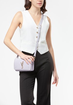 Donna con top bianco senza maniche con bottoni e pantaloni neri, che porta una piccola borsa a tracolla viola chiaro testurizzata con logo dorato.