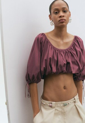 Kvinde iført en lilla crop-top med pufærmer og beige bukser med høj talje og dekorativ kædebælte, stående mod en hvid væg.