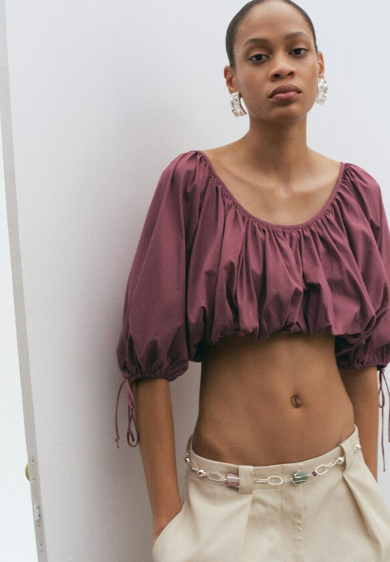 Femme portant un crop top violet à manches bouffantes, un pantalon taille haute beige avec une ceinture chaîne décorative, debout contre un mur blanc.