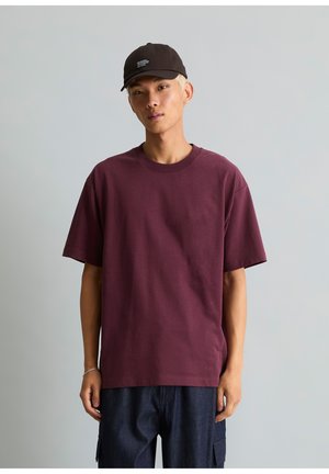 Jonge man die een donkerpaars oversized T-shirt, donkere cargobroek, zwarte pet en een zilveren armband draagt, staand voor een effen grijze achtergrond.
