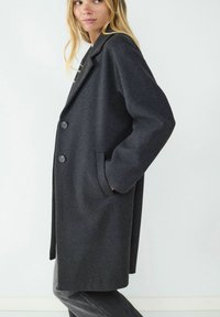 Grauer Wollmantel mit taillierter Passform, ausgestattet mit einem Reverskragen, zwei Knöpfen vorne und seitlichen Taschen. Glatte Textur.