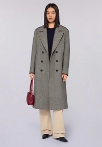 Grauer Wollmantel mit doppelreihigem Schnitt, breiten Revers, schwarzen Knöpfen und Seitentaschen, getragen über beigen Hosen und eine rote Tasche haltend.