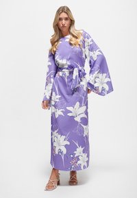 Langes, lila Blumenkleid mit großen weißen Lilienmotiven, weiten �Ärmeln und gebundenem Taillenbund. Glattes Gewebe, knöchellanges Design, kombiniert mit Riemchensandalen.