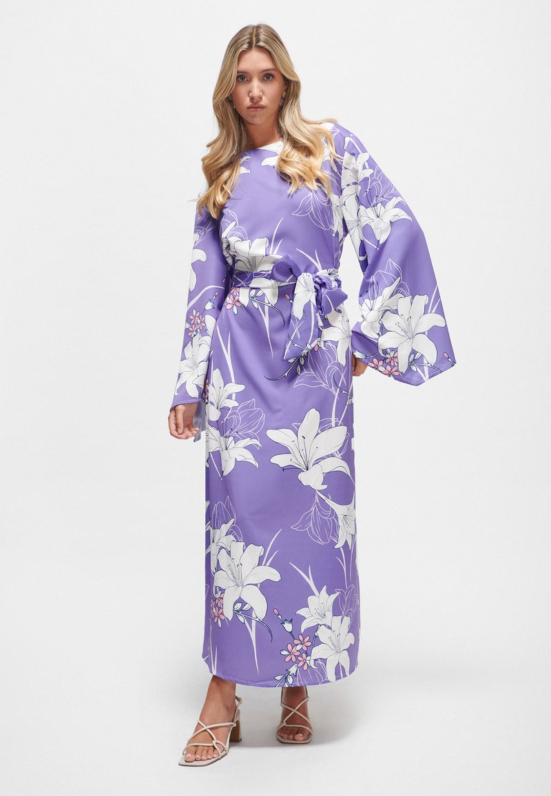 Langes, lila Blumenkleid mit großen weißen Lilienmotiven, weiten Ärmeln und gebundenem Taillenbund. Glattes Gewebe, knöchellanges Design, kombiniert mit Riemchensandalen.