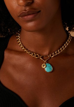Chunky gouden ketting met een grote turquoise hanger met zwarte aderen en een kleine accentcharme, liggend op een donkere stofachtergrond.