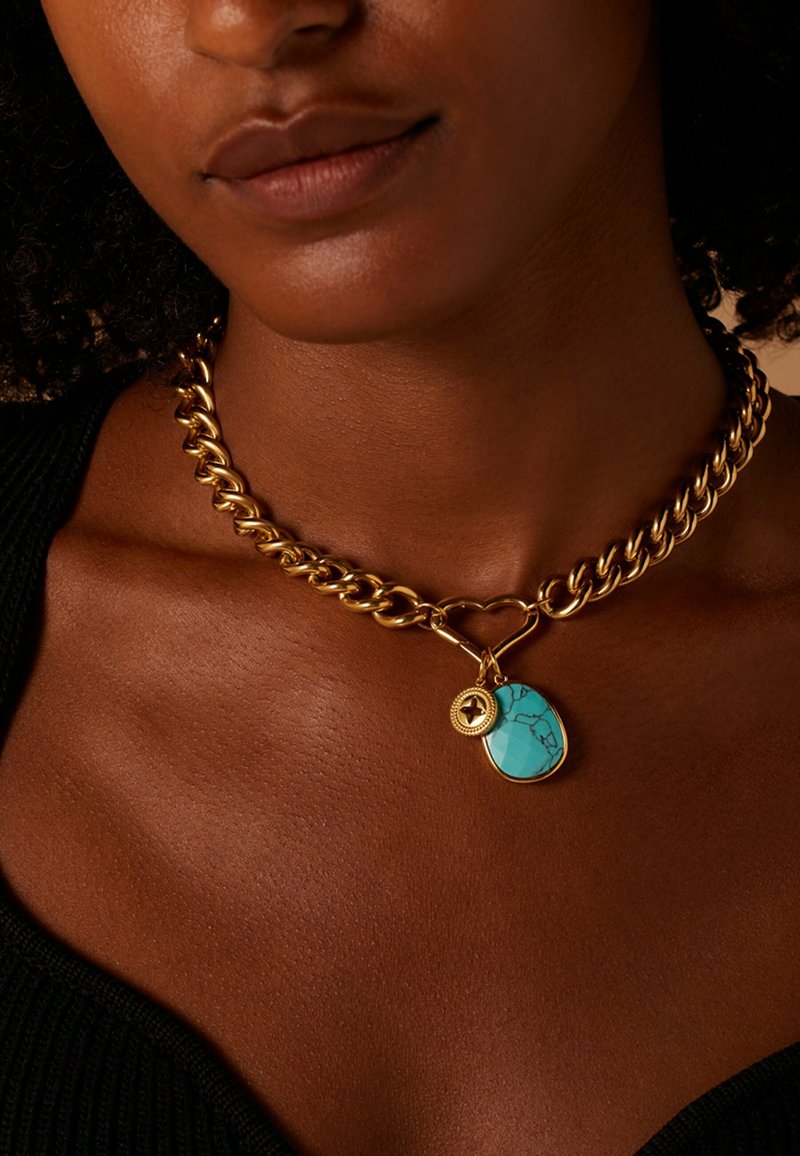 Chunky gouden ketting met een grote turquoise hanger met zwarte aderen en een kleine accentcharme, liggend op een donkere stofachtergrond.