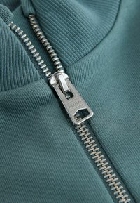 Mėlyna audinys su vingiuota tekstūra, viduryje esantis metalinis užtrauktukas. Artimas planas pabrėžia užtrauktuko dantų ir trauklės dizainą.