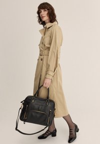 Gabardina beige con cintura ceñida, combinada con un bolso de hombro de cuero negro con herrajes dorados y múltiples cremalleras. Tacones negros brillantes.