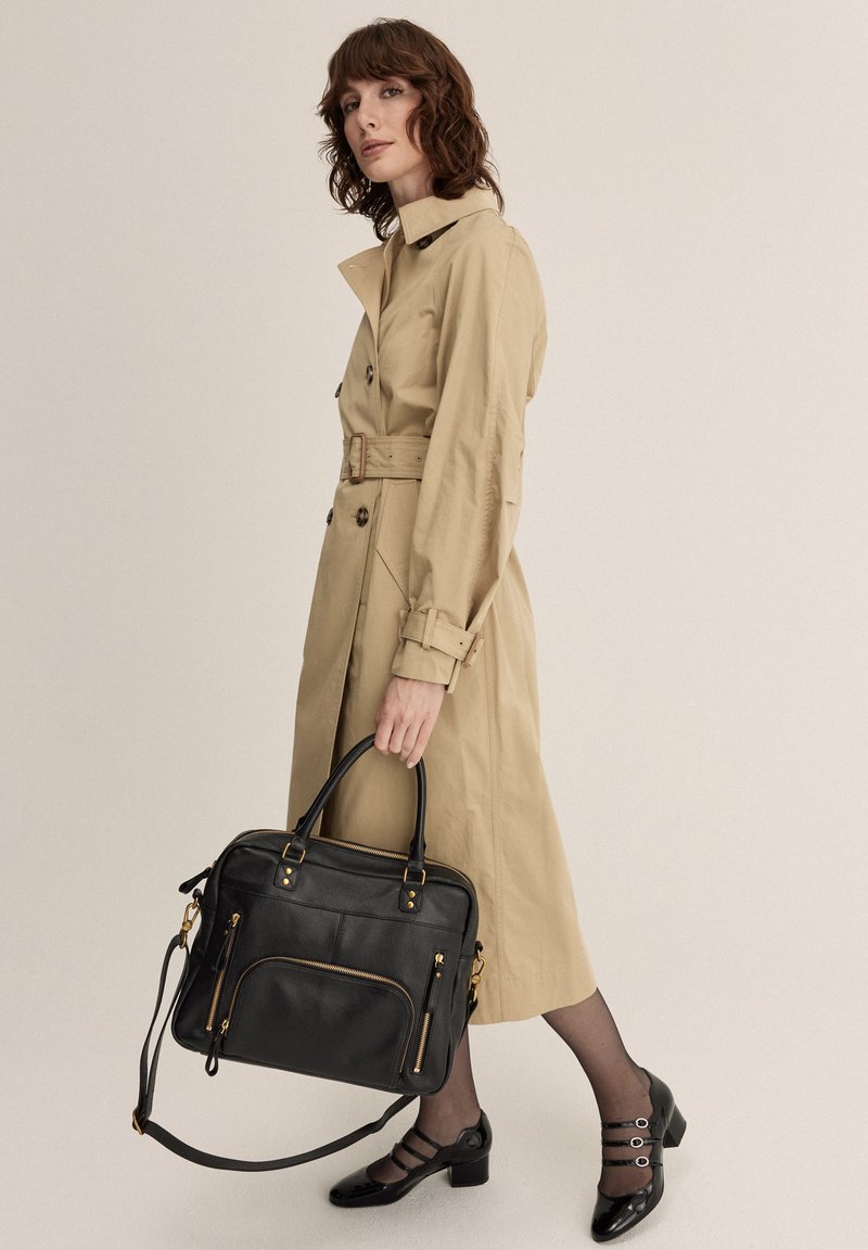 Trench coat beige con vita paperino, abbinato a una borsa di pelle nera con cerniere e dettagli dorati. Scarpe col tacco nere completano l'outfit.
