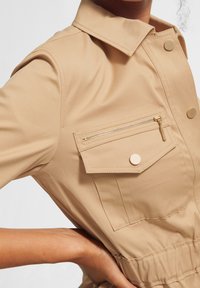 Primer plano de una chaqueta beige con cuello, bolsillo con cremallera, botones a presión y cintura elástica, llevada por una persona con la mano en la cadera.