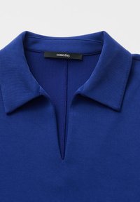 Chemise à col bleu avec une encolure en forme d'encoche en V et une étiquette noire visible portant la mention « someday. » sur un fond clair uni.