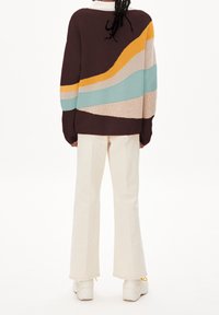 Pull en maille marron foncé avec des rayures ondulées en bleu clair, jaune et beige. Associé à un pantalon évasé crème et des baskets blanches.