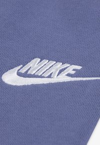 Logo Nike v bielom vyšívaní na textúrovanom fialovom látkovom pozadí. Dizajn obsahuje tvar swoosh s štýlizovaným písmom.