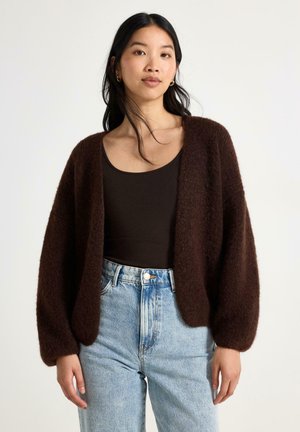 Donna che indossa un cardigan marrone scuro peloso sopra una maglietta nera e jeans azzurri a vita alta su uno sfondo semplice.